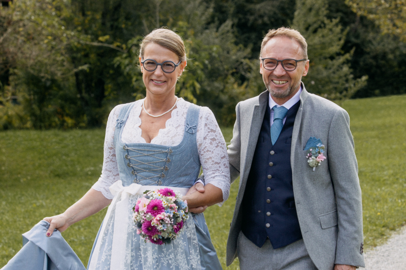 Brautpaar bei einer Allgäuer Hochzeit in moderner Tracht. Die Braut trägt ein hellblaues Dirndl mit Spitzenbluse, der Bräutigam einen modernen grauen Trachtenanzug mit Weste und Ansteckblume.